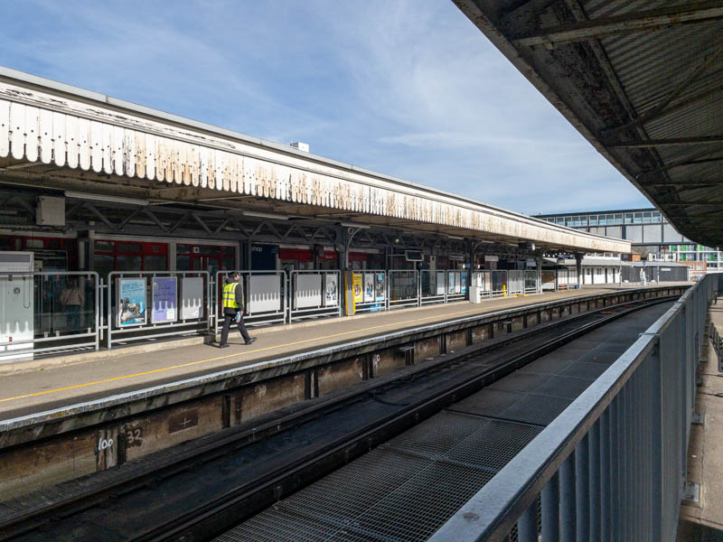 Platform 1 and its old
  wooden canopy from platform 3 opposite.  The platforms are gated off
  from the station entrance and facilities by a long fence, which has
  ticket barriers just at the very left of this picture.